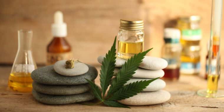 a photo of CBD oil in the foreground. There is a cannabis leaf resting next to it. In the background there are various nutritional supplements.